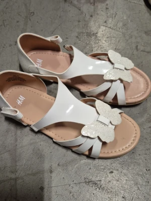 Sandaler - Snygga vita sandaler från H&M med glansig finish och dekorativa glitterfjärilar framtill. Sandalerna har öppen tå, justerbar rem runt hälen och mjuk beige innersula. Perfekta för sommarens alla tillfällen.