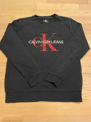 Svart sweatshirt Calvin Klein Jeans - Svart sweatshirt från Calvin Klein med stort rött och vitt CK-tryck på bröstet. Tröjan har rund halsringning, långa ärmar och ribbade muddar vid ärmslut och nederkant.