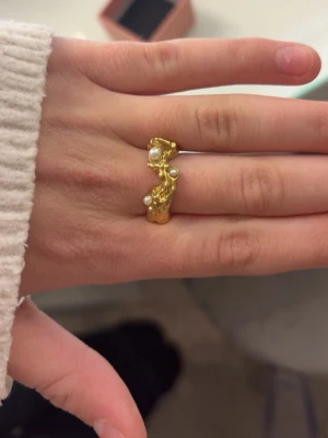 Guldfärgad ring med pärlor - Unik guldfärgad ring med organiskt, vågigt formspråk och små vita pärlor som dekoration. Ringen har en modern och konstnärlig design som sticker ut och passar dig som gillar statement-smycken.