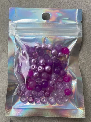 Lila och rosa pärlor i mixpåse - En mixpåse med pärlor i olika nyanser av lila och rosa. Pärlorna är runda och finns både som genomskinliga och pärlemorskimrande varianter. Perfekta till att skapa egna smycken eller accessoarer. Levereras i en holografisk zip-påse.