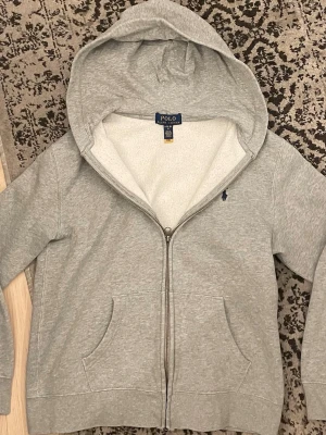 Grå zip hoodie från Polo Ralph Lauren - Klassisk grå hoodie från Polo Ralph Lauren med hel dragkedja, huva och två fickor fram. Broderad mörkblå logga på bröstet. Tillverkad i mjuk bomull för skön känsla och avslappnad look. Perfekt till jeans eller sweatpants. Tröjan har ett litet slitage på högra armen, men tröjan är använd ca 4-5 gånger.