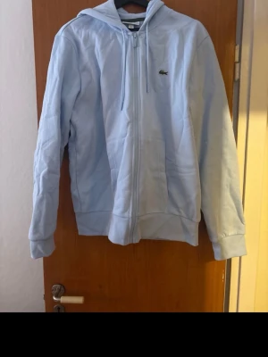Ljusblå hoodie från Lacoste - Snygg ljusblå hoodie från Lacoste med dragkedja framtill och klassisk krokodil-logga på bröstet. Modellen har huva med dragkedja och två stora fickor framtill. Tillverkad i mjukt bomullsmaterial för en skön känsla. Perfekt för en avslappnad och stilren look.