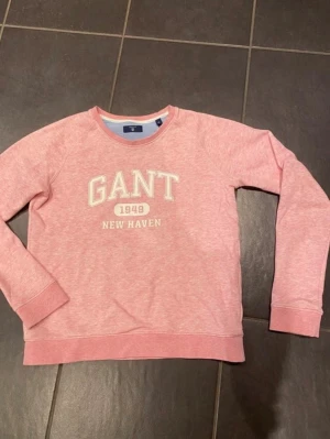 Rosa sweatshirt från GANT - Säljer en rosa sweatshirt från GANT med tryck 'GANT 1949 NEW HAVEN' på bröstet. Tröjan har rund halsringning, långa ärmar och ribbade muddar vid ärmslut och nederkant. Perfekt för en avslappnad och sportig stil.