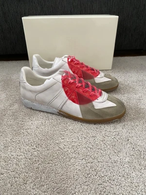 Maison Margiela Japan Gats - Unika sneakers från Maison Margiela i vitt och grått läder med beige mockadetaljer. Skorna har en stor röd sprayad hjärtformad detalj på sidan och matchande röda skosnören. Klassisk gummisula och låg profil för en clean look.
