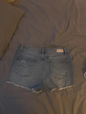 Blå jeansshorts med fransig kant - Säljer ett par klassiska blå jeansshorts med fransig rå kant och bakfickor. Shortsen har en ljus tvätt och en liten patch bak i midjan. Perfekta för sommaren och riktigt snygga till sneakers eller sandaler.