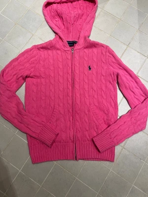 Rosa kabelstickad hoodie Ralph Lauren - Säljer en rosa kabelstickad hoodie från Ralph Lauren Sport i 100% bomull. Tröjan har dragkedja, huva, fickor framtill och den klassiska loggan broderad på bröstet. Perfekt för dig som gillar en sportig men ändå söt stil.