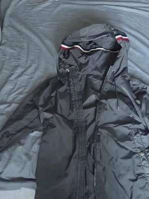 Svart vindjacka med huva - Snygg svart vindjacka med dragkedja och huva. Jackan har vita och röda detaljer på huvan samt justerbara snören. Tillverkad i ett glansigt, vindtätt material som passar perfekt för blåsiga dagar. Storlek 1 enligt etiketten.