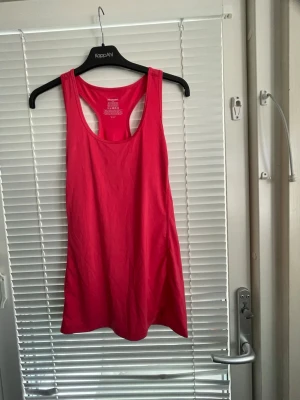 Rött träningslinne från Newbody - Snyggt rosa träningslinne från Newbody i funktionsmaterial. Linnet har brottarrygg och är gjort av 92% polyester och 8% elastan, vilket gör det stretchigt och bekvämt för träning. Perfekt för gymmet eller löprundan. Känns ej som XL utan mer som L