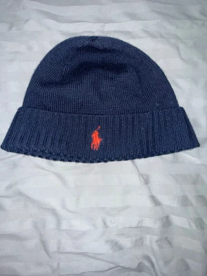 Mörkblå mössa från Polo Ralph Lauren - Snygg mörkblå mössa från Polo Ralph Lauren i mjuk merinoull. Klassisk ribbad kant och ikonisk röd logga broderad framtill. Perfekt accessoar för kalla dagar och enkel att matcha med olika outfits.