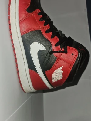 Air Jordan 1 High röd/svart/vit - Säljer ett par Air Jordan 1 High sneakers från Nike i klassisk röd, svart och vit färg. Skorna har en vit swoosh på sidan, svart snörning och den ikoniska Air Jordan-loggan på ankeln. Tillverkade i läder med en vit mellansula och röd yttersula.