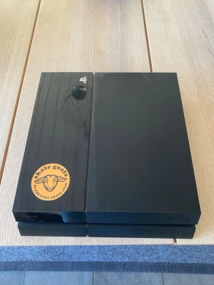 Sony PlayStation 4 svart konsol - Säljer en svart PlayStation 4 konsol från Sony. Konsolen har en stilren, rektangulär design med matta och blanka ytor samt en sticker på ovansidan. Perfekt för gaming och streaming, med två USB-portar på framsidan. Kommer med en USB sladd och en strömsladd