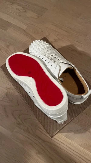 Vita Louboutin sneakers med nitar - Snygga vita sneakers från Christian Louboutin med klassisk röd sula och coola nitar på tån. Skorna är låga och har vita skosnören samt detaljer i vitt läder. Perfekta för dig som vill sticka ut med exklusiv design.