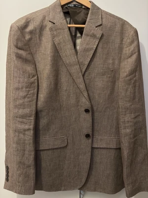 Brun kavaj i linne från Selected Homme - Snygg brun kavaj från Selected Homme i ett svalt linnematerial. Klassisk modell med två knappar framtill, smala slag och tre fickor. Perfekt för en stilren look med en modern touch.