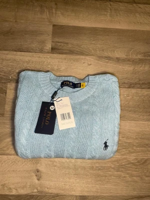 Ljusblå kabelstickad tröja Polo Ralph Lauren - Ljusblå kabelstickad tröja från Polo Ralph Lauren med rund halsringning och klassisk broderad logga på bröstet. Tröjan har långa ärmar och ribbade muddar vid ärmslut och nederkant. Tillverkad i mjukt material för en skön känsla.