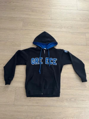 Svart hoodie med GREECE-tryck - Snygg svart hoodie med dragkedja och blå insida i huvan. På bröstet står det 'GREECE' i stora vita och blå bokstäver. Huvan har blå snören och på ärmen finns en liten grekisk flagga. Perfekt för dig som gillar streetwear och vill sticka ut.