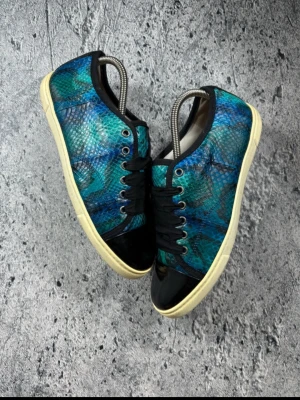 Snakeskin lanvins - Unika sneakers med blå-grönt ormmönster, svarta detaljer och vit sula. Skorna har svarta snören och glansiga partier vid tå och häl. Perfekt för dig som vill sticka ut med färg och mönster. Materialet är en mix av textil och syntet. Bara skorna ingår och ingen innersula