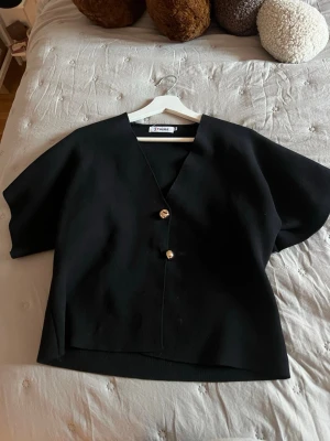 Svart v-ringad topp med knappar - Stilren svart topp med v-ringning och två dekorativa guldfärgade knappar framtill. Toppen har korta ärmar och en rak, något boxig passform. Perfekt att matcha med både jeans och kjol för en trendig look.