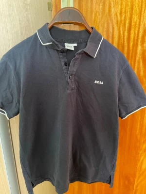 Svart pikétröja från BOSS - Klassisk blå  pikétröja från BOSS med vit piping på krage och ärmslut. Tröjan har korta ärmar, knappar framtill och BOSS-logga broderad på bröstet. Materialet är bekvämt och bra kvalitet. Fint skick inte använd särskilt mycket.Det står att de är en 16m men den är ganska liten skulle jag säga.Passar personer som är cirkus 150cm skulle jag säga. 