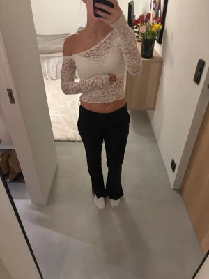 Vit spets offshoulder croppad topp - Säljer en vit croppad topp i spets med offshoulder och lång ärm. Toppen har en snygg asymmetrisk design med ena axeln bar och är tillverkad i ett tunt, stretchigt spetsmaterial. Tröjan är genomskinlig, men på bilden har jag valt att täcka upp med ett linne under🫶🏻