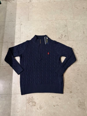 Mörkblå stickad tröja Polo Ralph Lauren - Mörkblå kabelstickad tröja från Polo Ralph Lauren med halv dragkedja och röd broderad logga på bröstet. Tröjan har ribbade muddar och hög krage, perfekt för en klassisk och stilren look. 