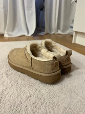 Beige fårskinnstofflor från UGG - Mysiga beige skor från UGG med mjukt fårskinnsfoder och mockaliknande utsida. Tofflorna har en rejäl sula i gummi och en låg slip-in modell med liten hälflik. Perfekta för att hålla fötterna varma.