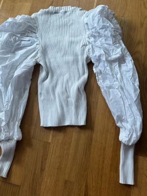 Vit ribbad topp med puffärmar - Trendig vit topp med ribbad kropp och stora puffärmar i kontrasterande tyg. Ärmarna är långa och avslutas med breda ribbade muddar. Perfekt för dig som vill sticka ut med en cool och unik look.