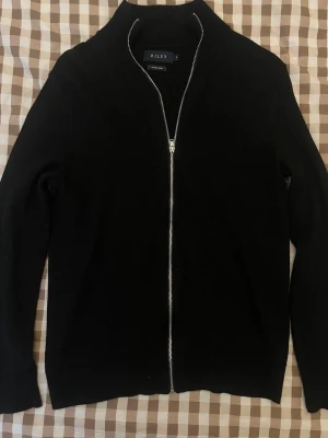 Svart merinoullskofta från Riley - Stilren svart kofta från Riley i mjuk merinoull. Koftan har hel dragkedja framtill, hög krage och långa ärmar. Perfekt för lager på lager och enkel att matcha med olika outfits. Tröjan har inga skador och är använt max 3-4 gånger. Ordinarie pris 1199kr