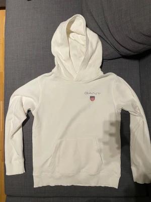 Vit hoodie från GANT med logga - Säljer en stilren vit hoodie från GANT med broderad logga och sköld på bröstet. Hoodien har stor huva och en klassisk magficka. Tillverkad i mjukt material som känns skönt mot huden. Perfekt för dig som gillar enkel och clean stil. Pris går att diskuteras.