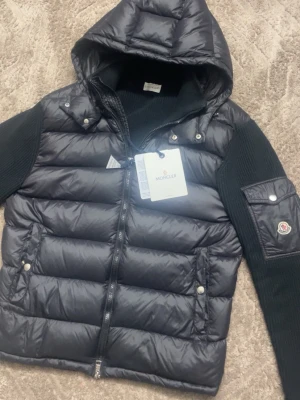 Svart dunväst med huva från Moncler - Snygg svart dunväst från Moncler med stickade svarta ärmar och huva. Västen har två framfickor med tryckknappar, dragkedja framtill och en Moncler-logga på ärmen. Materialet är glansigt och quiltat, perfekt för en trendig streetwear-look.