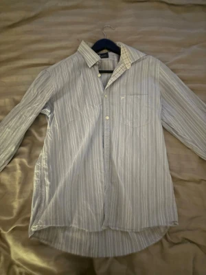 Ljusblå randig skjorta från peakperformance - Säljer en ljusblå skjorta från peakperformance med tunna vita ränder och klassisk krage. Skjortan har lång ärm, bröstficka och knappar framtill. Manschetterna har ett rutigt mönster på insidan för en extra detalj. Perfekt för en clean och stilren look.