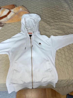 Vit hoodie med dragkedja och huva - Snygg vit hoodie från Moose Knuckles med dragkedja framtill och två fickor. Klassisk huva med dragsko och diskret logga på bröstet. Perfekt för en avslappnad streetwear-look. Tillverkad i mjukt bomullsmaterial.