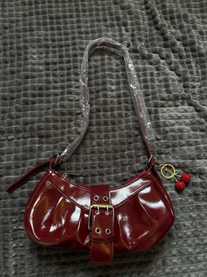 Vinröd axelväska med körsbärsberlock - Snygg oanvänd vinröd handväska i glansigt konstläder med justerbar axelrem och dekorativt spänne framtill. Väskan har en söt körsbärsberlock i rött och grönt som detalj. Perfekt för dig som gillar trendiga accessoarer med lite extra attityd.
