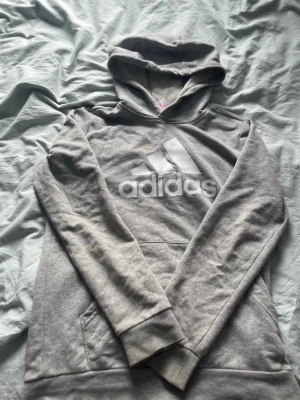 Grå hoodie från Adidas med logga - Snygg grå hoodie från Adidas med stor vit logga på bröstet. Klassisk huva och känguruficka framtill. Tillverkad i mjukt bomullsmaterial som känns skönt mot huden. Perfekt för en avslappnad och sportig stil. Priset kan även diskuteras om de behövs 👌