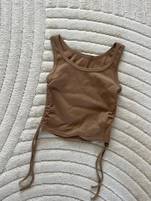 Brun ribbad linnetopp med snörning - Snygg brun ribbad linnetopp med snörning i sidorna. Toppen är ärmlös och croppad, perfekt för varma dagar eller att styla lager på lager. Mjuk och stretchig känsla i materialet.