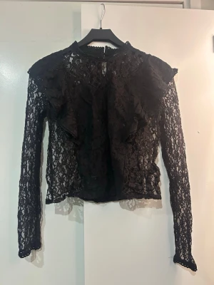 Svart spetsblus med volanger - Snygg svart blus i spets med volangdetaljer framtill och vid axlarna. Endast använd 1 gång🌟