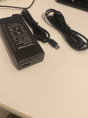  Elscoter laddare  - Säljer en svart power adapter till dator med tillhörande kablar. Märkt med 'Power Charger' och har tydliga specifikationer på ovansidan. Passar till olika typer av elektronik som kräver extern strömförsörjning. Tillverkad i plast och har rund kontakt.