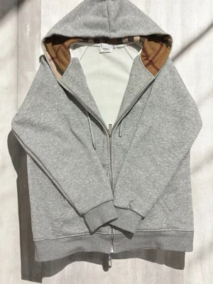 Grå zip-hoodie från Burberry - Snygg grå hoodie med dragkedja från Burberry. Klassisk huva med det ikoniska rutmönstret på insidan, ribbade muddar och fickor framtill. Perfekt för en chill och stilren look. Materialet är mjukt och bekvämt, passar till det mesta.