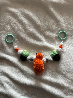 Barnvagnshänge med dinosaurie 🧶🦕 - Ett virkat barnvagnshänge med massor av charm ⭐️  Designat med en orange dinosaurie med blåa taggar, söta kaktusar samt vita och grå bollar - lekfullt men harmoniskt i färgerna.   Mått: 📏 cirka 45 cm lång från ring till ring. Med rundresår så den är elastisk   Detaljer:   - elastiskt snöre - smidigt att fästa och ger lite följsamhet  - perfekt för barnvagn, babyskydd eller babygym  - stimulerar barnets nyfikenhet med färg och form.  