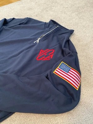 Jeffrey Epstein Quarter Zip - Mörkblå Jeffrey Epstein Island tröja med halv dragkedja och ståkrage. Röd broderad logga på bröstet och en stor amerikansk flagga på ena ärmen. Tröjan har ribbade muddar vid ärmslut och nederkant. Materialet ser ut att vara mjukt och bekvämt, perfekt för en avslappnad stil.