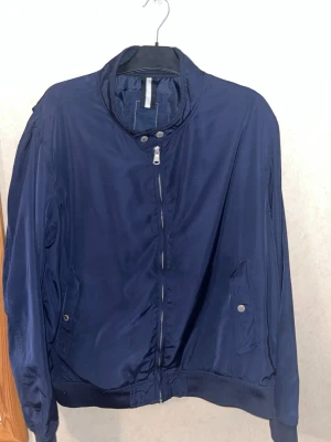 Mörkblå bomberjacka med dragkedja - Snygg mörkblå bomberjacka med klassisk ribbad mudd i ärmslut och nederkant. Jackan har snedställda fickor med tryckknappar och en hög krage med knappstängning. Tillverkad i ett glansigt, tunt material som ger en modern look.