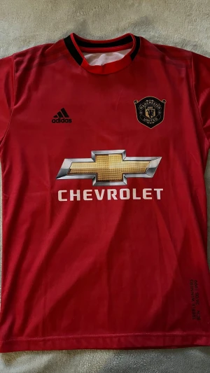 Manchester United Pogba matchtröja - Röd Manchester United matchtröja från Adidas med Pogba och nummer 6 på ryggen. Tröjan har klubbmärke, Adidas-logga och Chevrolet som sponsor framtill. Svart och röd krage, tillverkad i lätt och ventilerande material. Perfekt för fotboll eller som supporterplagg.