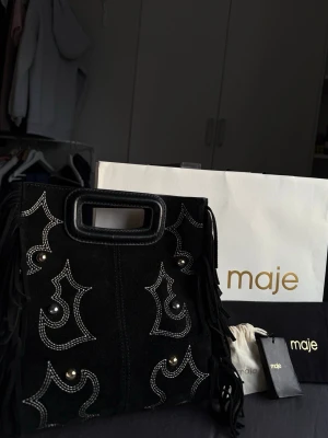  Maje väska i mocka  - Snygg svart handväska i mocka från Maje med dekorativa silvernitar, vita sömmar och fransar på sidorna. Väskan har ett fyrkantigt handtag och medföljande axelrem. Perfekt för dig som vill ha en unik accessoar med coola detaljer. Den stora modellen och är limited edition så går inte att köpa längre!!