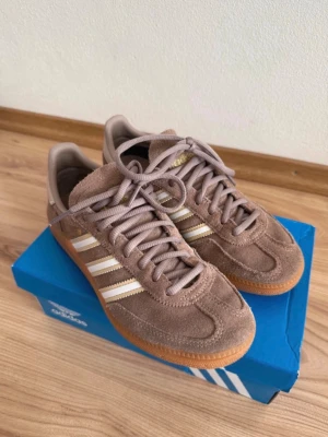 Bruna Adidas spezial sneakers - Säljer ett par bruna Adidas Spezial sneakers i mocka med klassiska vita och guldiga stripes på sidan. Skorna har gummisula och lågt skaft, samt matchande bruna skosnören. Perfekta för dig som gillar retrostil och streetwear.