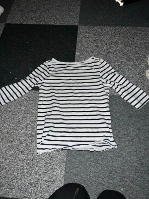 Randig topp från Esmara med knappar - Snygg vit topp från Esmara med mörkblå horisontella ränder och trekvartsärm. Toppen har dekorativa knappar på ena axeln och är gjord i mjuk bomull. Perfekt för en avslappnad och stilren look. Stl M men passar S