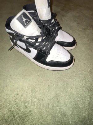 Svarta och vita Air Jordan 1 Mid - Säljer ett par klassiska Air Jordan 1 Mid i svart och vitt läder med svarta snören och ikoniska loggor på sidan och plösen. Snygg kontrast mellan färgerna och tidlös design som alltid är rätt. Perfekta för dig som gillar streetwear och sneakerskultur men tyvärr har jag inte box 📦.
