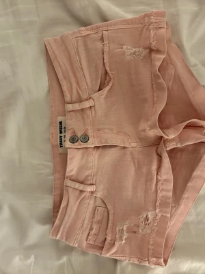 Ljusrosa shorts från Tally Weijl - Supersnygga lågmidjade ljusrosa shorts från Tally Weijl med  slitna detaljer. Klassisk femficksmodell med två knappar framtill och bälteshällor. Perfekta för varma dagar och ger en avslappnad vibe. Midjemått 40