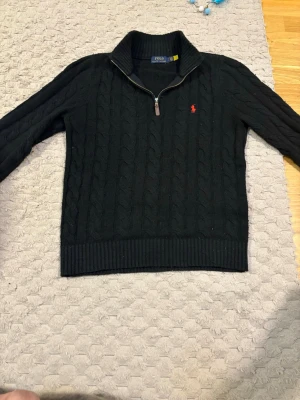 Svart kabelstickad tröja Polo Ralph Lauren - Kabelstickad svart tröja från Polo Ralph Lauren med halv dragkedja och hög krage. Tröjan har ett rött broderat logomärke på bröstet och ribbade muddar vid ärmslut och nederkant fast den är nopprig. Perfekt för lager på lager och klassisk stil. 