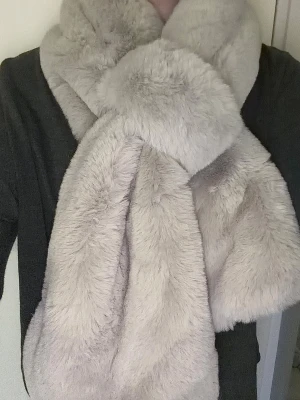 Grå halsduk K.zell - Halsduken har en smart "nyckelhålsöppning" som gör att du enkelt kan trä den ena änden genom den andra, vilket gör att den sitter perfekt på plats.  Skick: Mycket gott skick Färg: Ljusgrå Material: Mjuk fuskpäls (faux fur) 