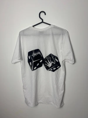 Vit Stüssy t-shirt med tärningsmotiv - Säljer en vit t-shirt från Stüssy med svart tryck av två tärningar på både bröstet och ryggen. T-shirten har rund hals, korta ärmar och är gjord i mjuk bomull. Perfekt för dig som gillar streetwear och vill ha en clean men ändå unik look.