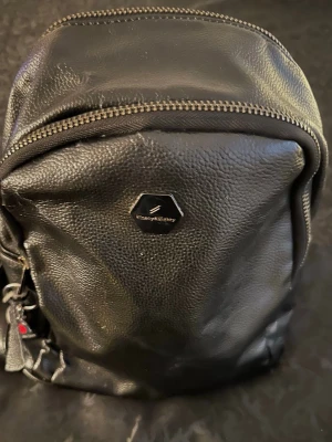 Svart ryggsäck från Everyday Kitchy - Snygg svart ryggsäck i konstläder från Everyday Kitchy. Väskan har flera fack med dragkedja, justerbara axelband och en stilren hexagonlogga framtill. Perfekt storlek för att bära med sig det viktigaste och enkel att matcha till olika outfits.